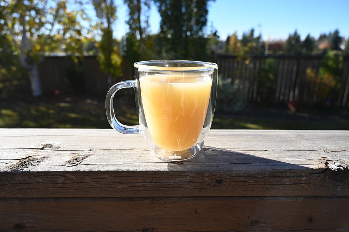 Image of a clear mug with bone broth in it on a deck outside ready to sip. There are trees in the background.
