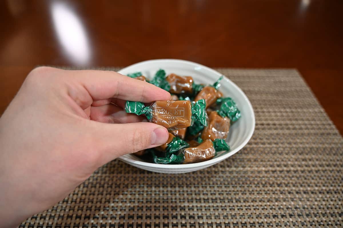Closeup image of a hand holding one wrapped caramel, in the background is a bowl full of wrapped caramels.