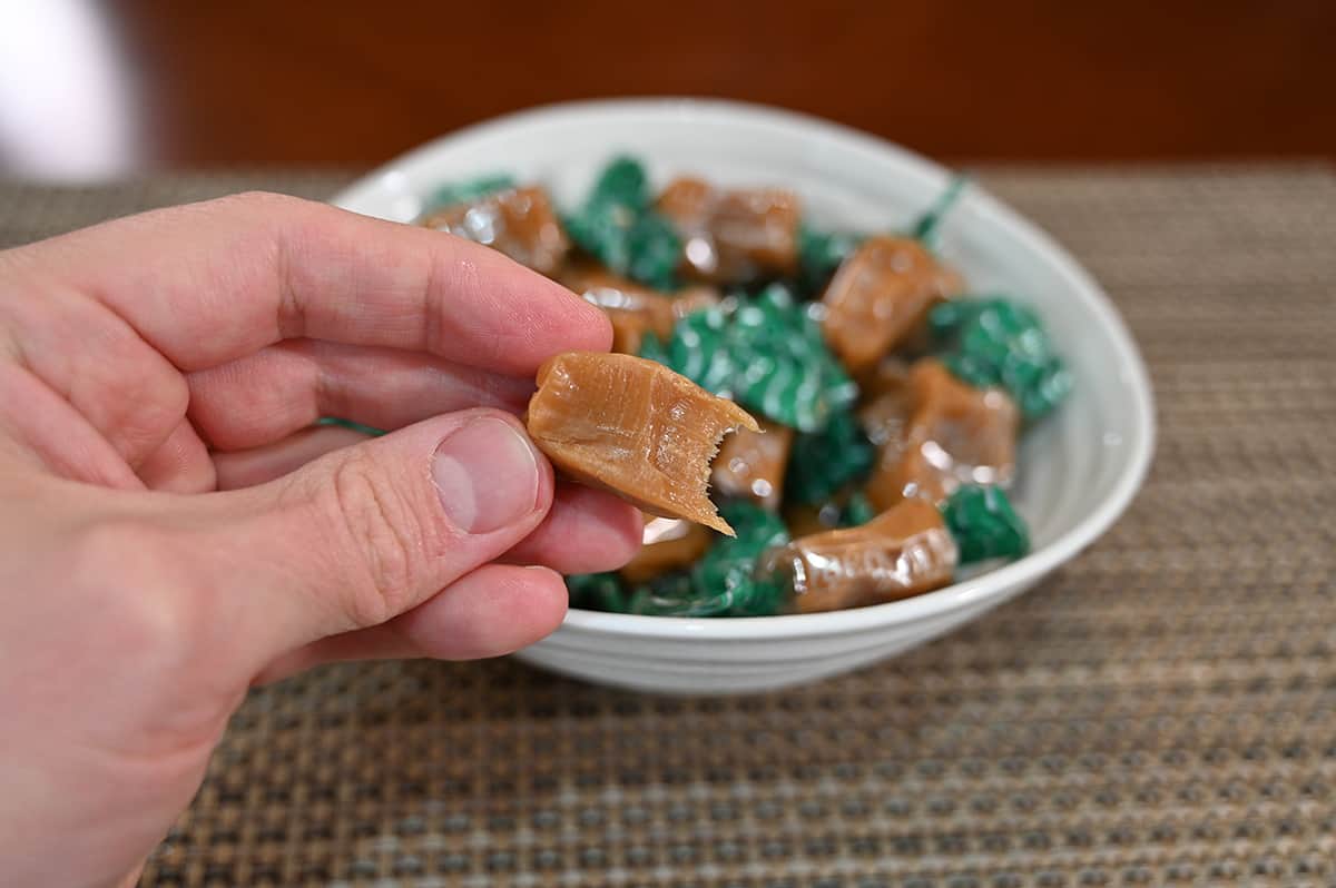 Closeup image of a hand holding one caramel with a bite taken out of it, in the background is a bowl full of wrapped caramels.