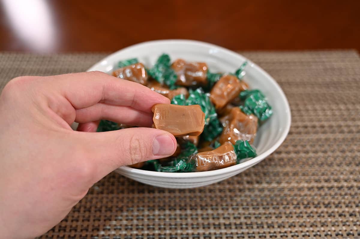 Closeup image of a hand holding one caramel unwrapped, in the background is a bowl full of wrapped caramels.