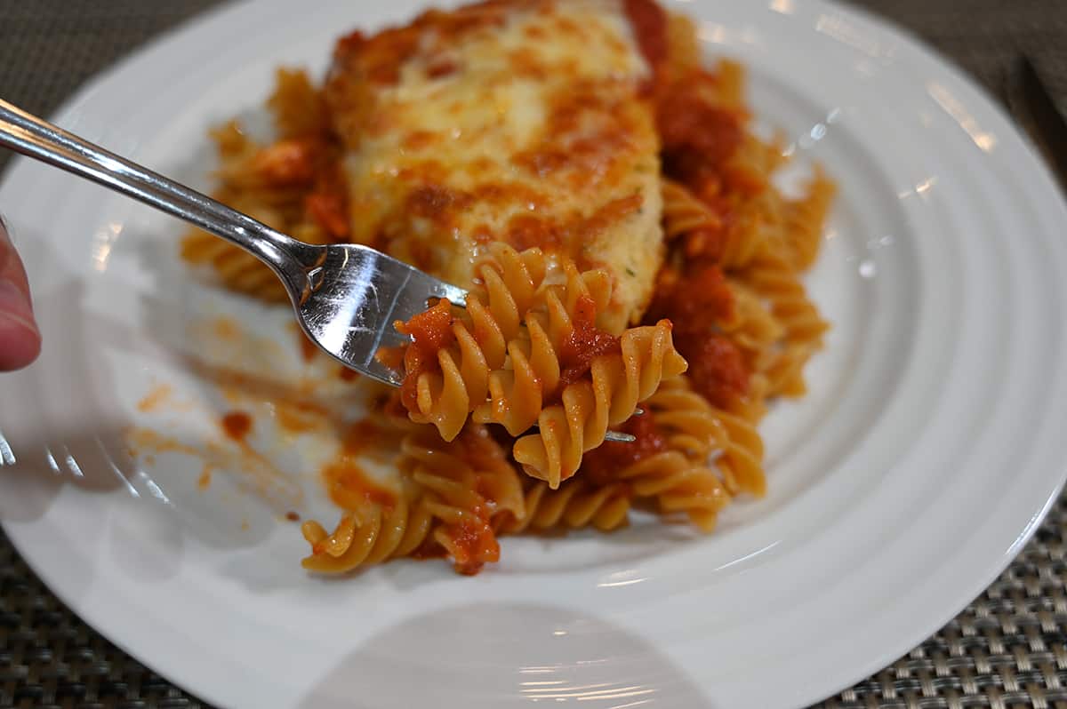 Closeup image of a fork with chickpea pasta on it close to the camera, in the background of the image is pasta and chicken parmesan on a white plate.