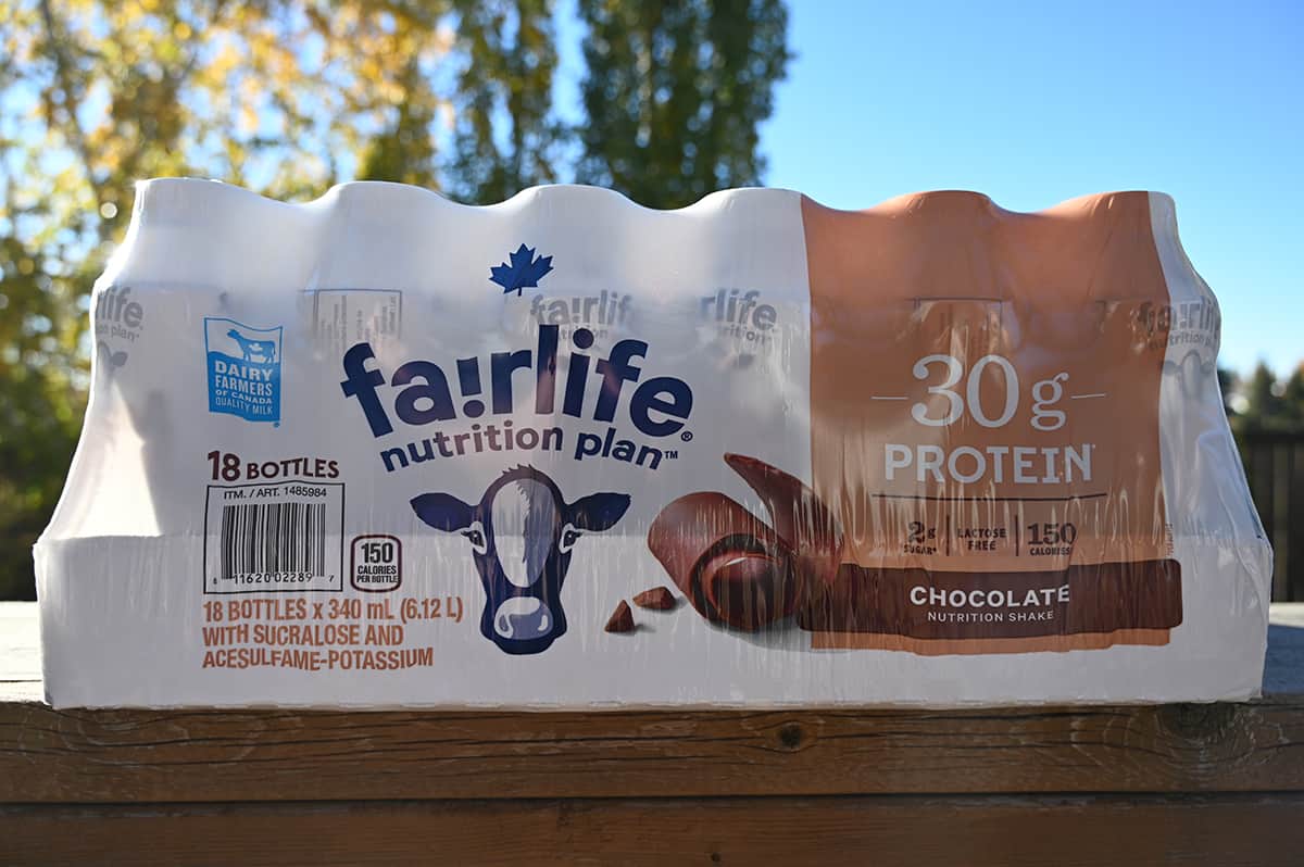 Image of a case of chocolate Fairlife protein shakes sitting on a deck outside with trees and a blue sky in the background.