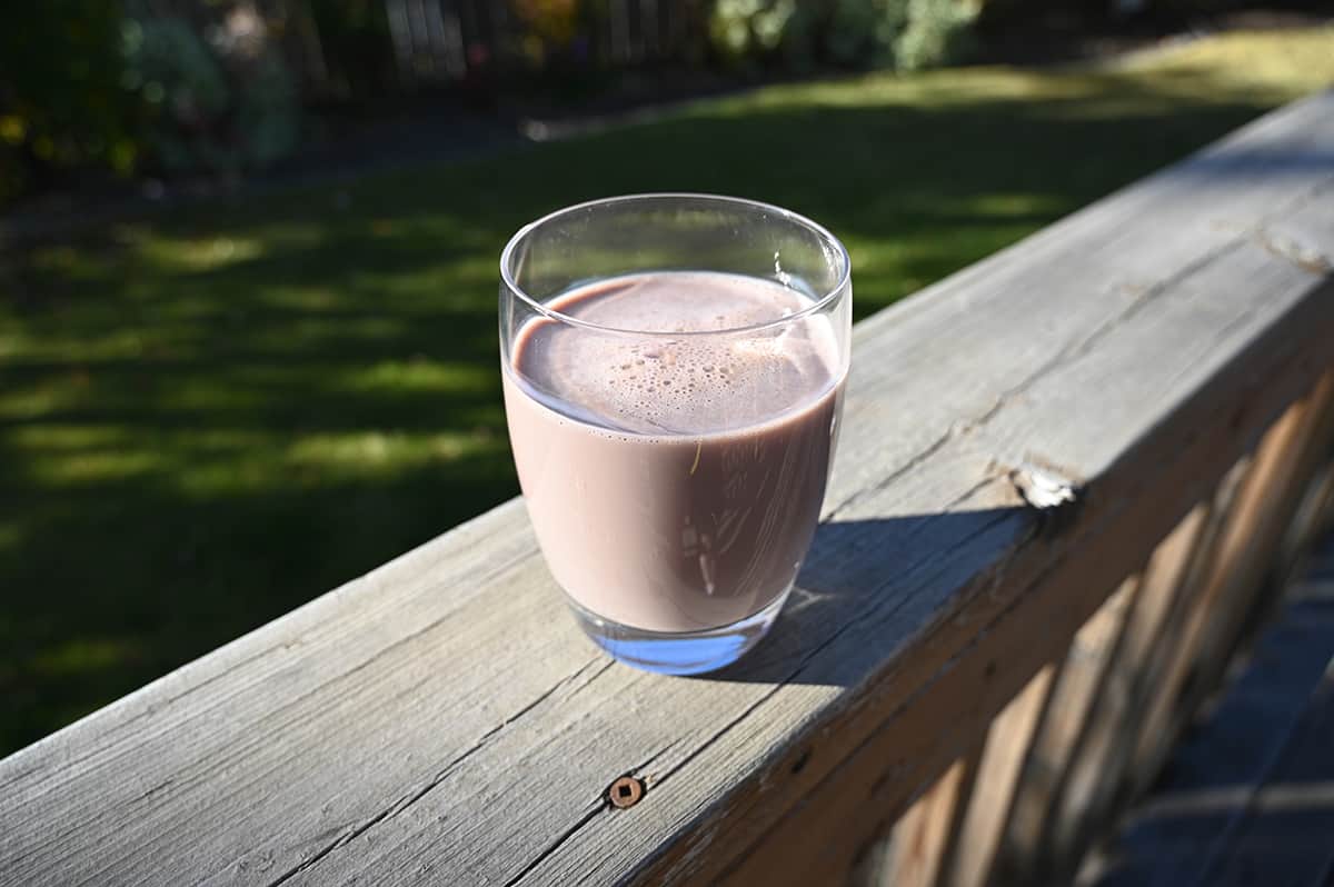 Image of a clear glass with chocolate protein shake poured into it. The glass is sitting on a deck.