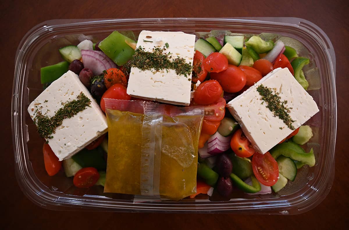 Top down image of the Greek salad opened and sitting on a table. The image shows three large blocks of feta sitting on top of the salad and one large packet of dressing also on top.