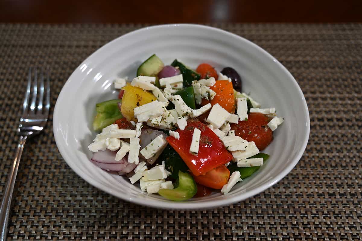 Sideview image of the Greek salad served in a white bowl with crumbled feta on top. There are large chunks of peppers, olives, herbs, cucumbers and tomatoes visible.