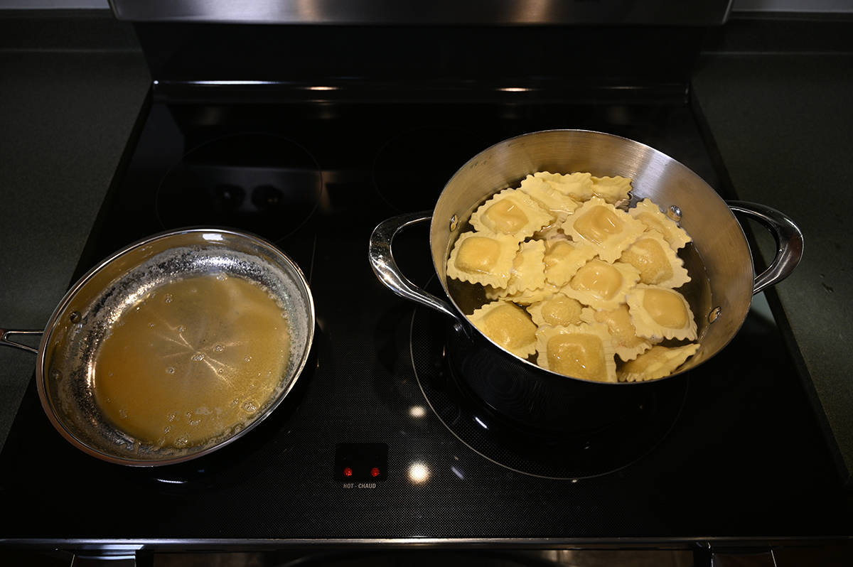 Top down image of a pot of boiling water with ravioli being cooked inside next to a fry pan with garlic butter sauce on a stovetop.