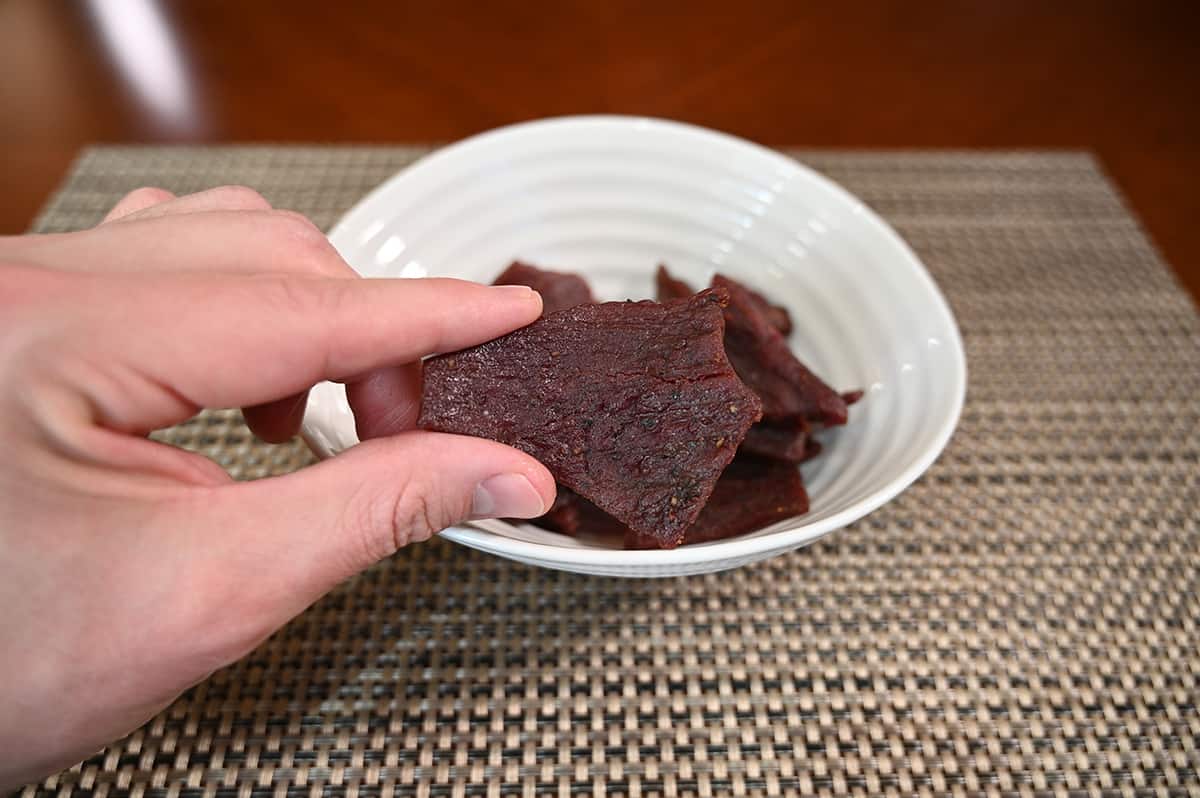 Closeup image of a hand holding a piece of jerky close to the camera. In the background of the image is a bowl of jerky.