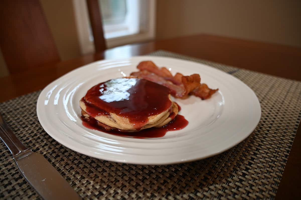 Side view image of a stack of pancakes with fruit syrup drizzled on top beside two pieces of bacon, served on a white plate.