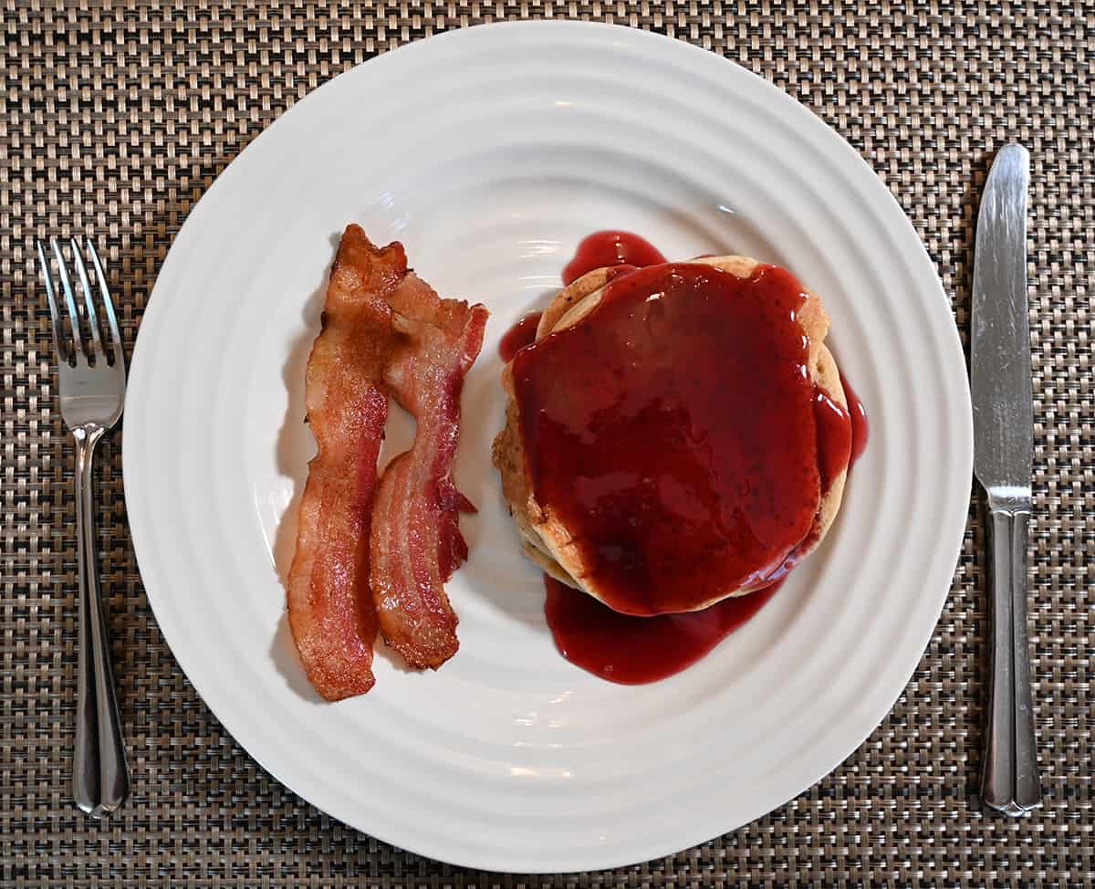 Top down image of a stack of pancakes with fruit syrup drizzled on top beside two pieces of bacon, served on a white plate.