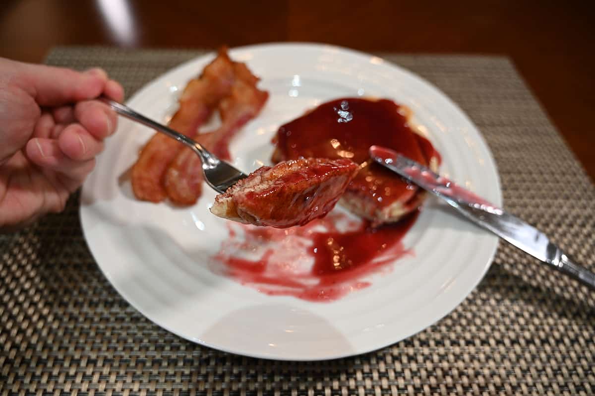 Side view closeup image of a plate with pancakes and bacon on it, close to the camera is a fork with a piece of pancake drizzled with fruit syrup.