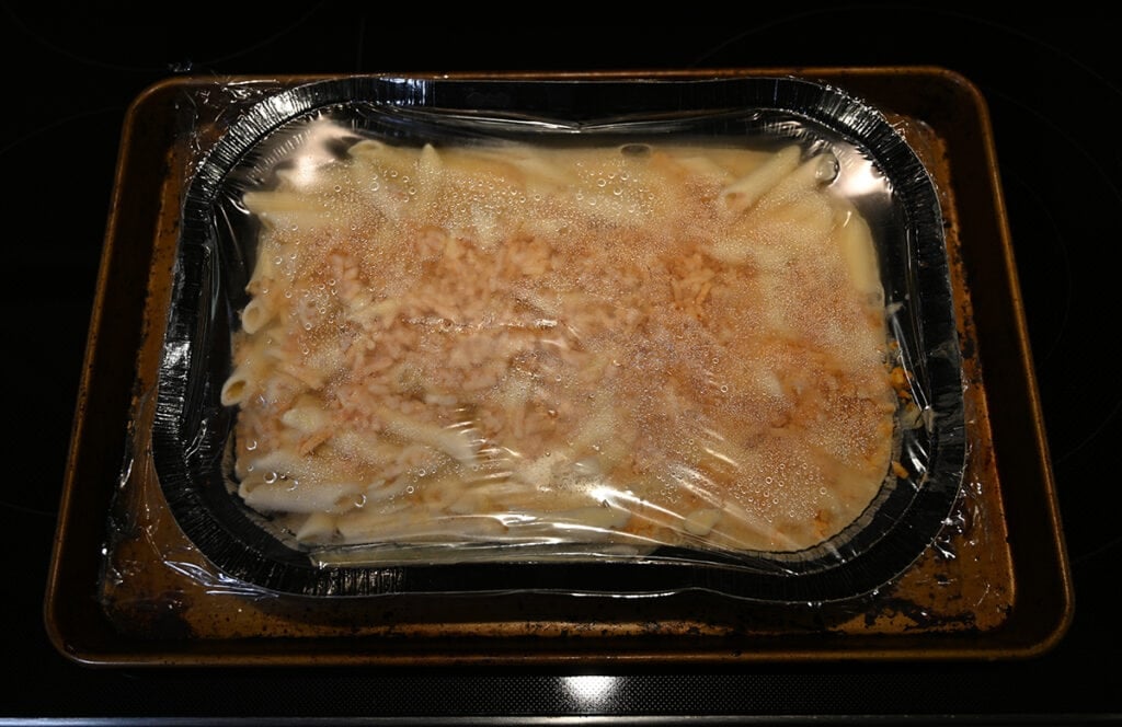 Top down image of the frozen tray of mac and cheese sitting on a baking tray. There has been a slit cut in the plastic film on top.