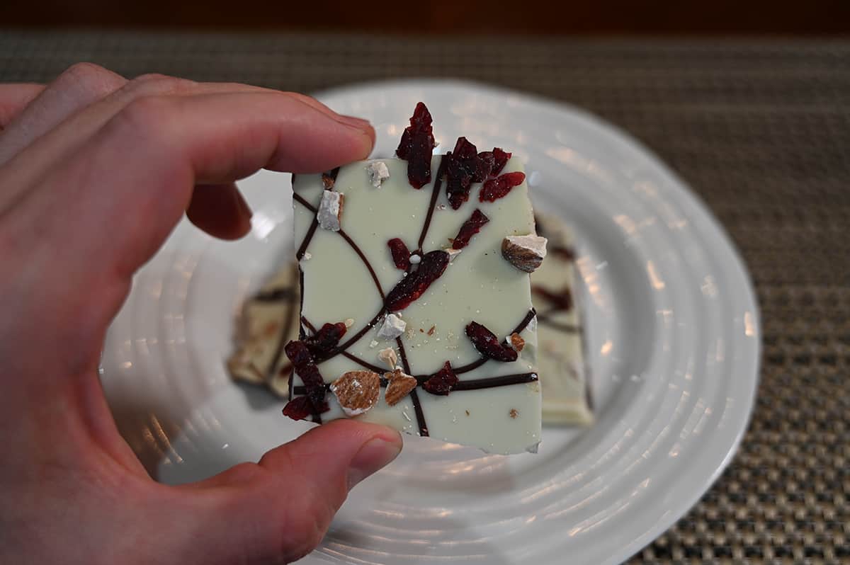 Closeup image of a hand holding a piece of cranberry almond bark close to the camera so you can see the almonds and cranberries on the bark.