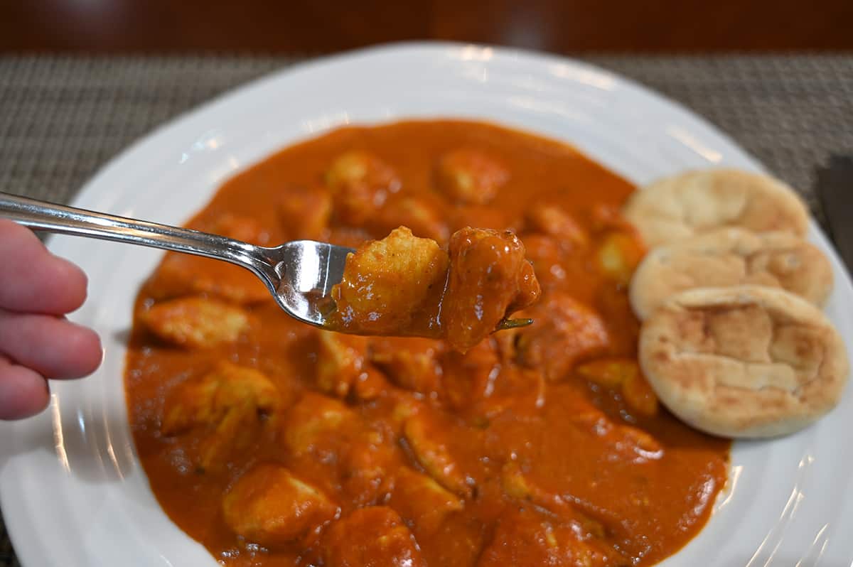 Closeup image of a fork with chicken tikka masala on it with a plate of it in the background beside naan bread.