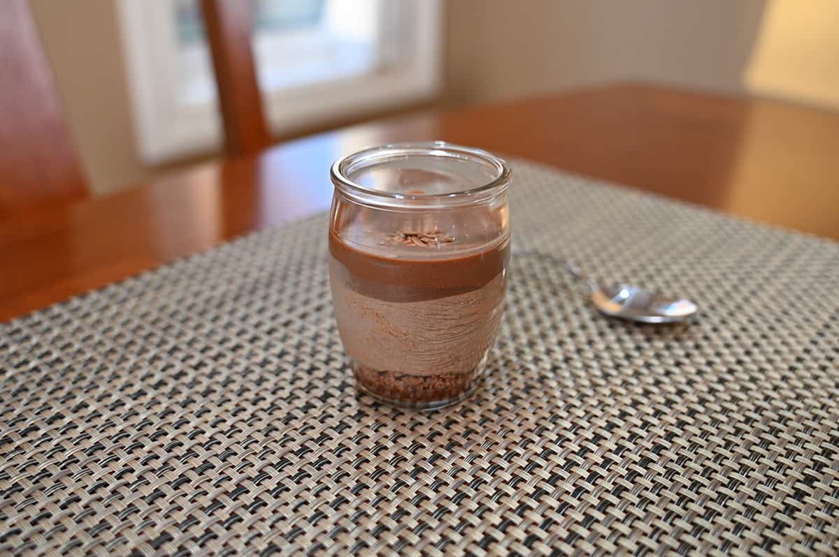 Sideview image of one chocolate mousse cup sitting on a table beside a spoon, unopened.