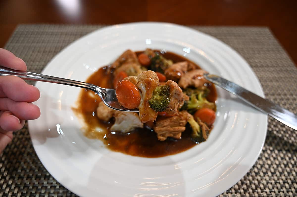 Closeup side view image of a hand holding a fork with chicken stir-fry on it. In the background is a plate of chicken stir-fry.