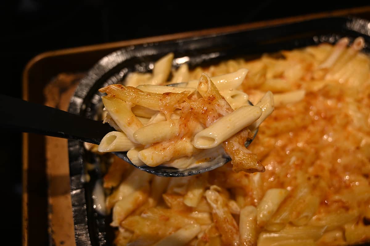Closeup image of a serving spoon, serving mac and cheese out of tray after being baked in the oven. There are crispy top noodles visible and softer bottom noodles.