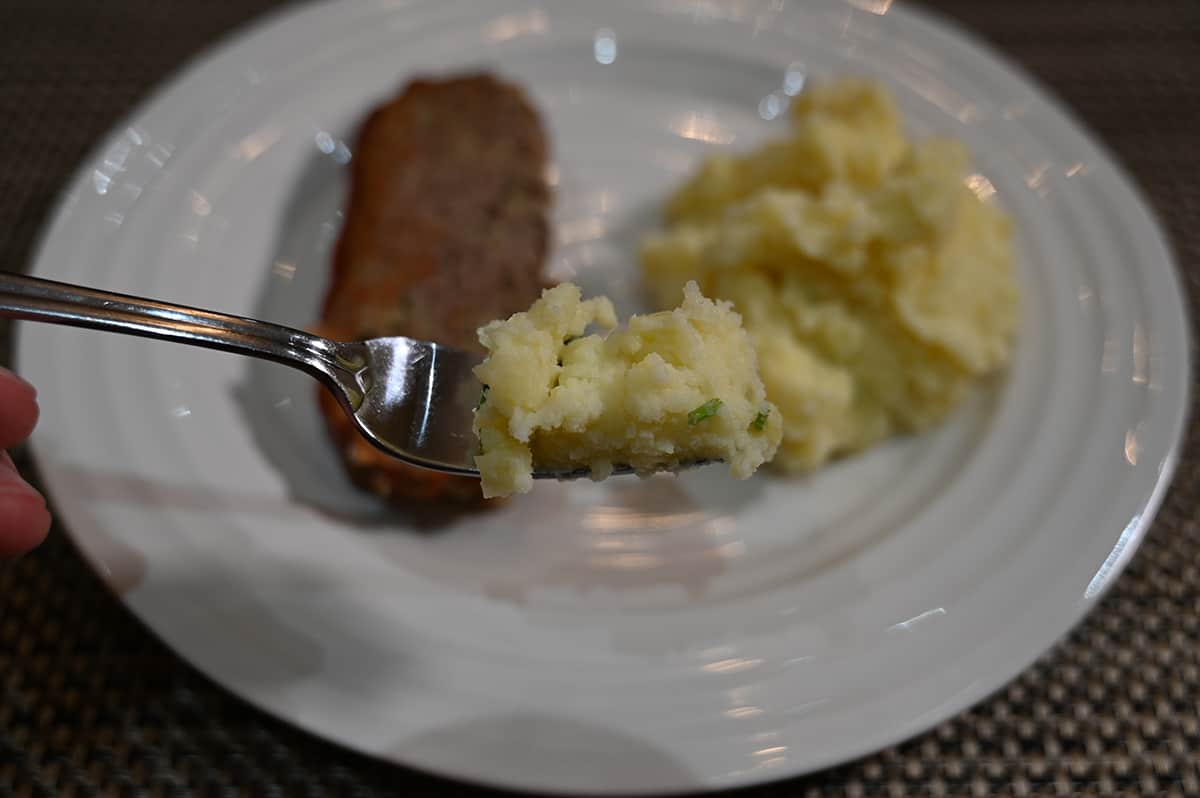 Closeup image of a forkful of mashed potatoes. In the background is mashed potatoes and meatloaf on a plate.