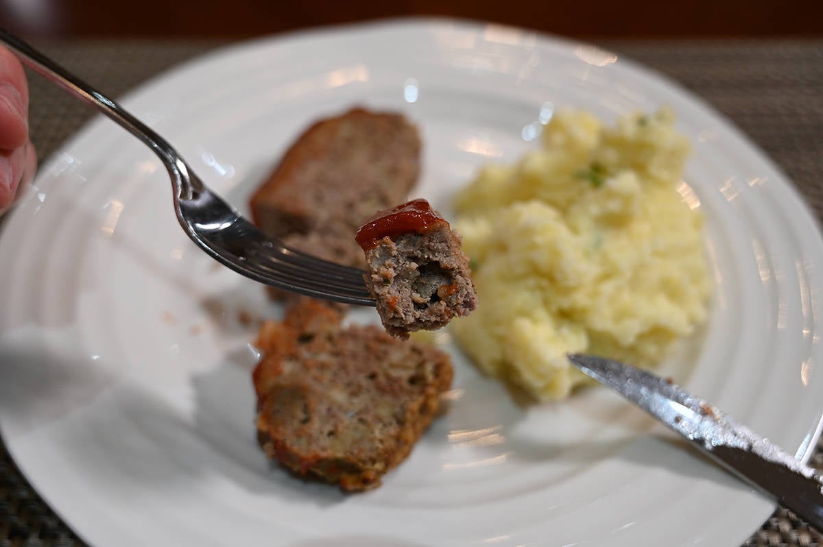 Closeup image of a forkful of meatloaf. In the background is mashed potatoes and meatloaf on a plate.