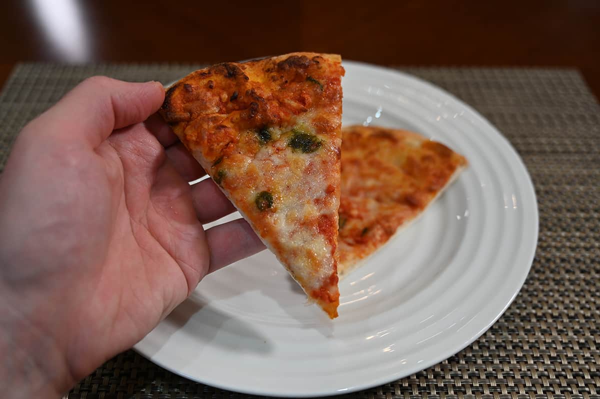 Closeup image of a hand holding one slice of pizza close to the camera with another slice in the background on a white plate.