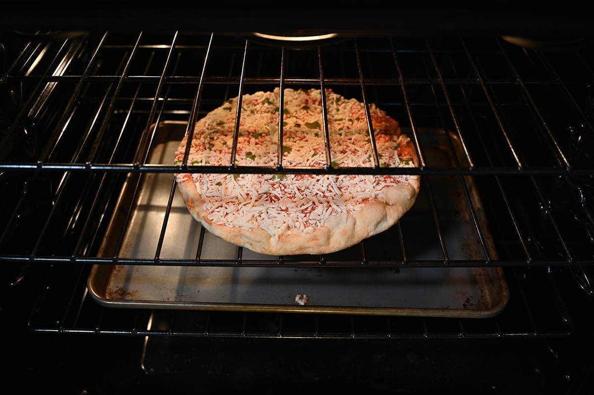Side view image of a frozen pizza baking in an oven directly on the middle rack with a cookie tray underneath on the bottom rack.