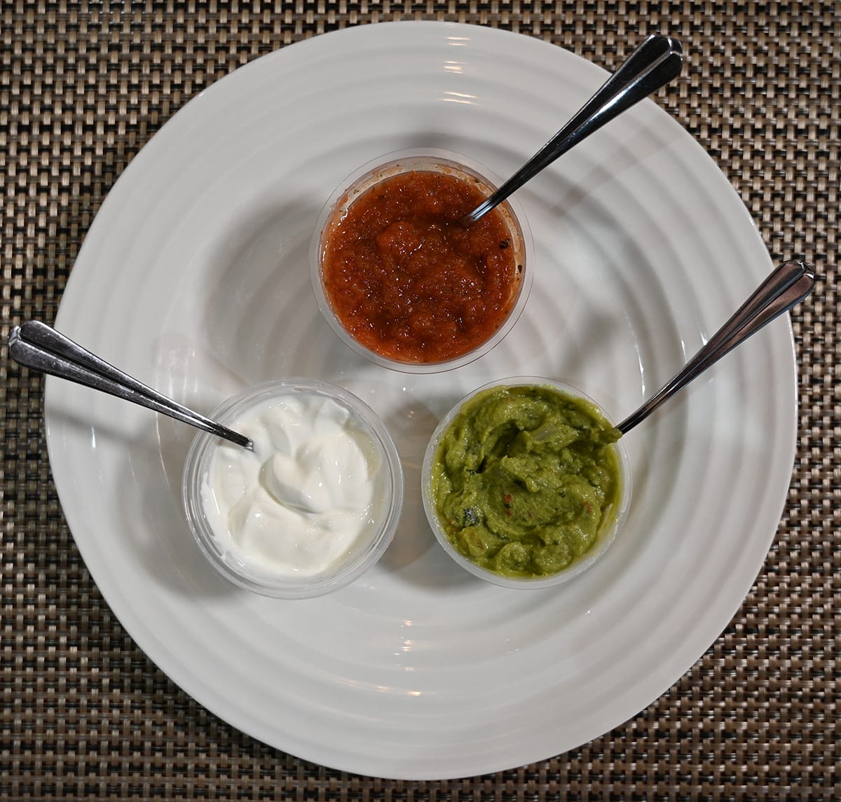 Top down image of the salsa, sour cream and guacamole containers sitting on a white plate with spoons in them.
