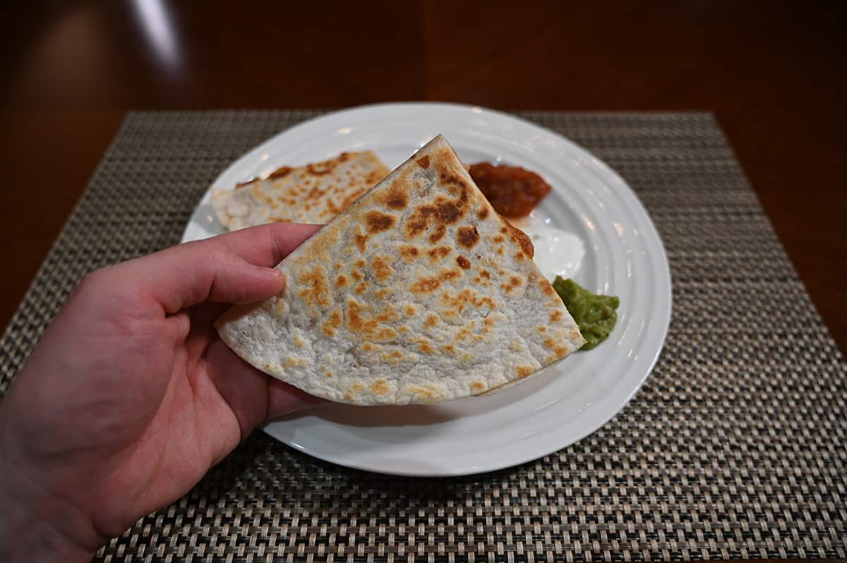 Side view image of a hand holding one quesdilla close to the camera, in the background is a plate with salsa, sour cream and guacamole.