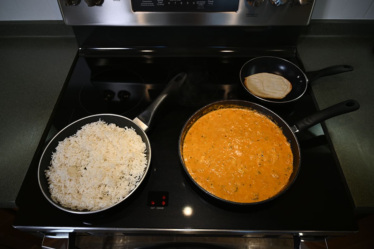 Top down image of a stovetop with a pan of rice, a pan of curry and a pan with naan cooking on top.