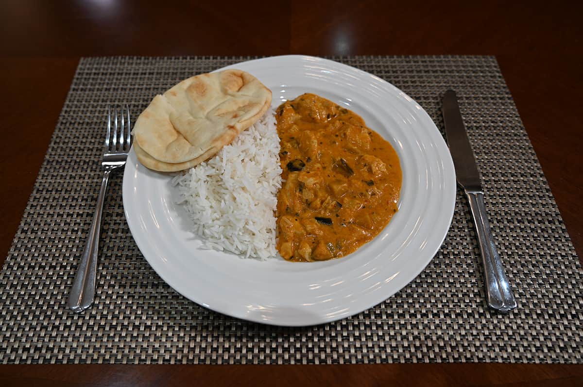 Side view image of a plate loaded with curry, rice and naan served on a table beside a knife and fork.
