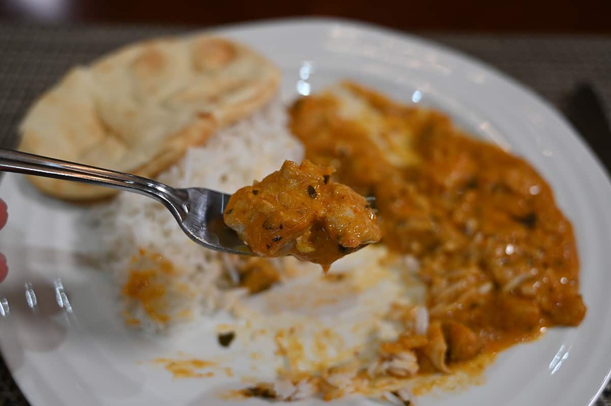 Closeup image of a fork with chicken curry on it, in the background of the image if a plate with rice, curry and naan on it. 