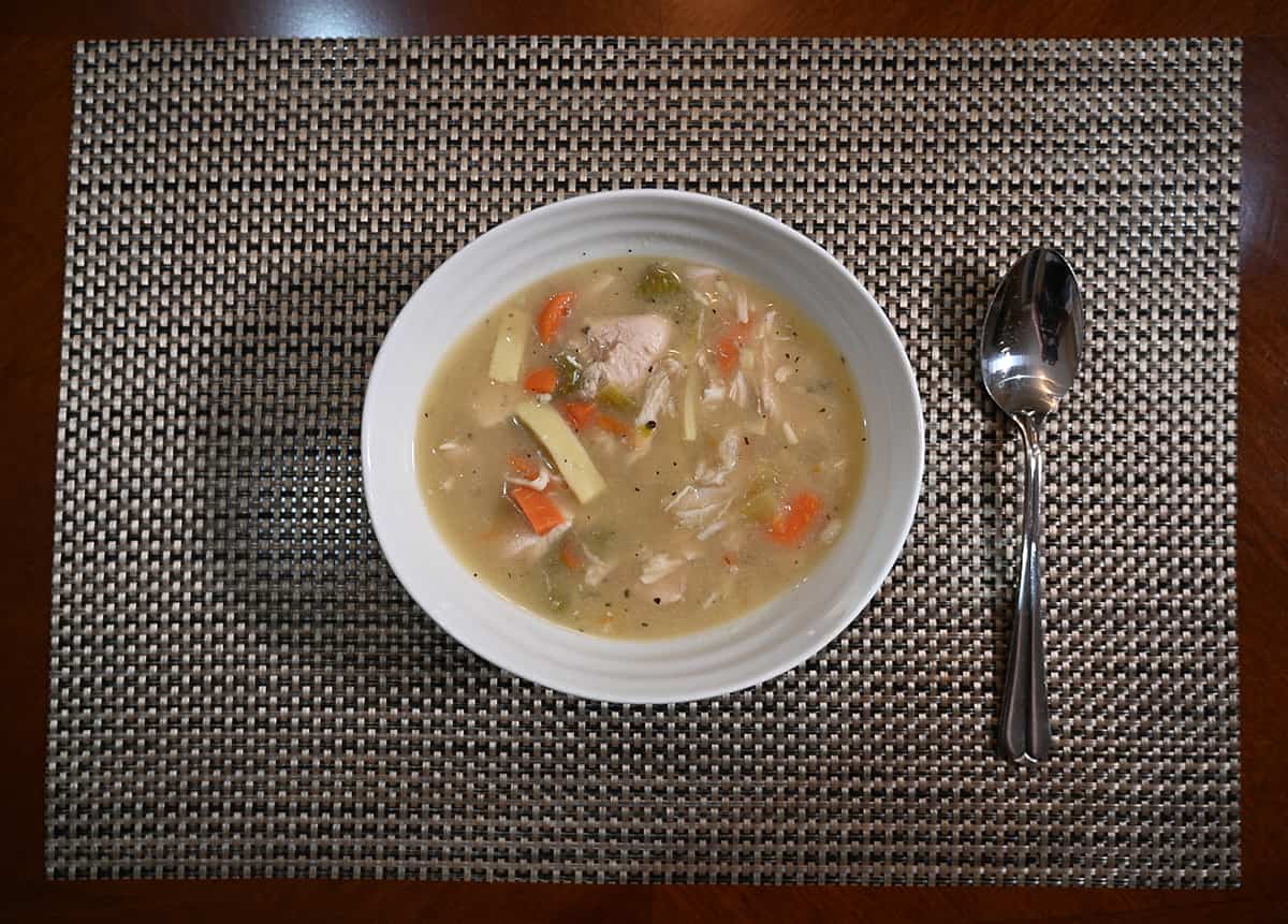 Top down image of a bowl of chicken noodle soup beside a spoon on a table.