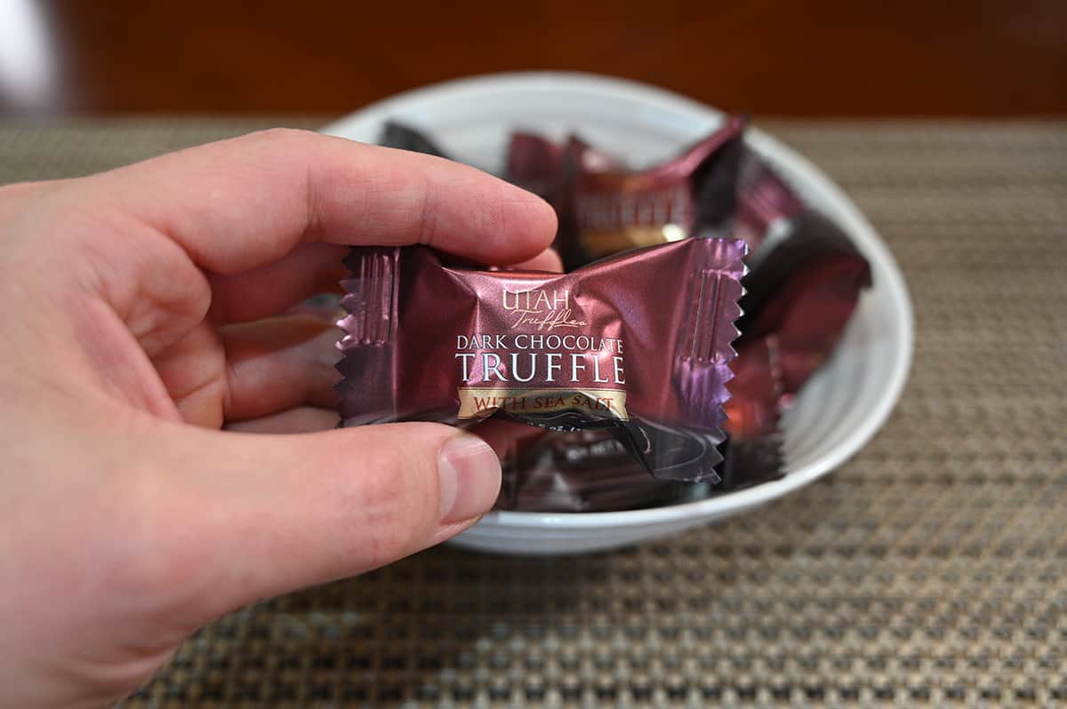 Top down image of a bowl full of individually wrapped dark chocolate with sea salt truffles with a hand holding one individually wrapped truffle.