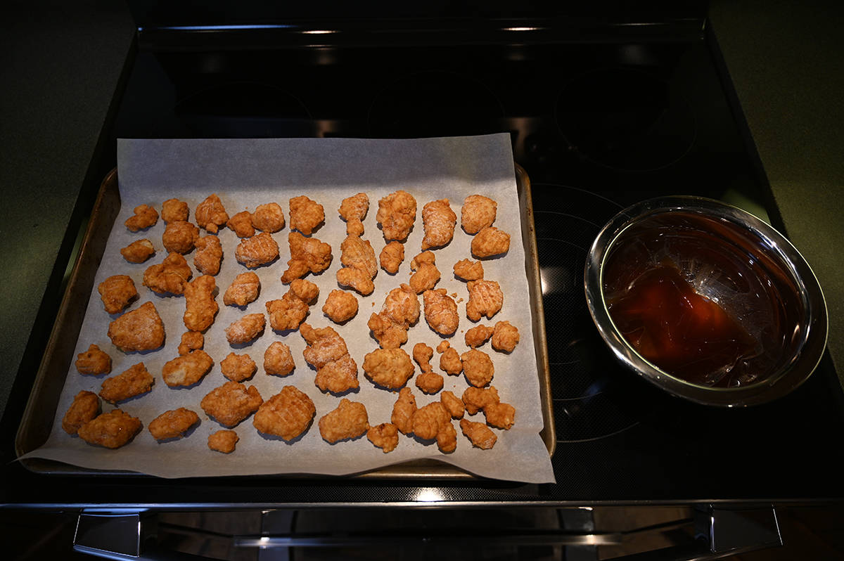 Top down image of chicken pieces spread out of a parchment lined baking sheet beside a bowl wth a sauce packet warming in water.
