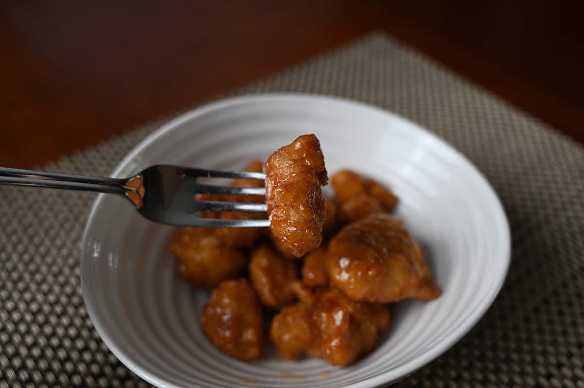 Closeup image of a fork holding a piece of orange chicken close to the camera with a bowl of orange chicken in the background.