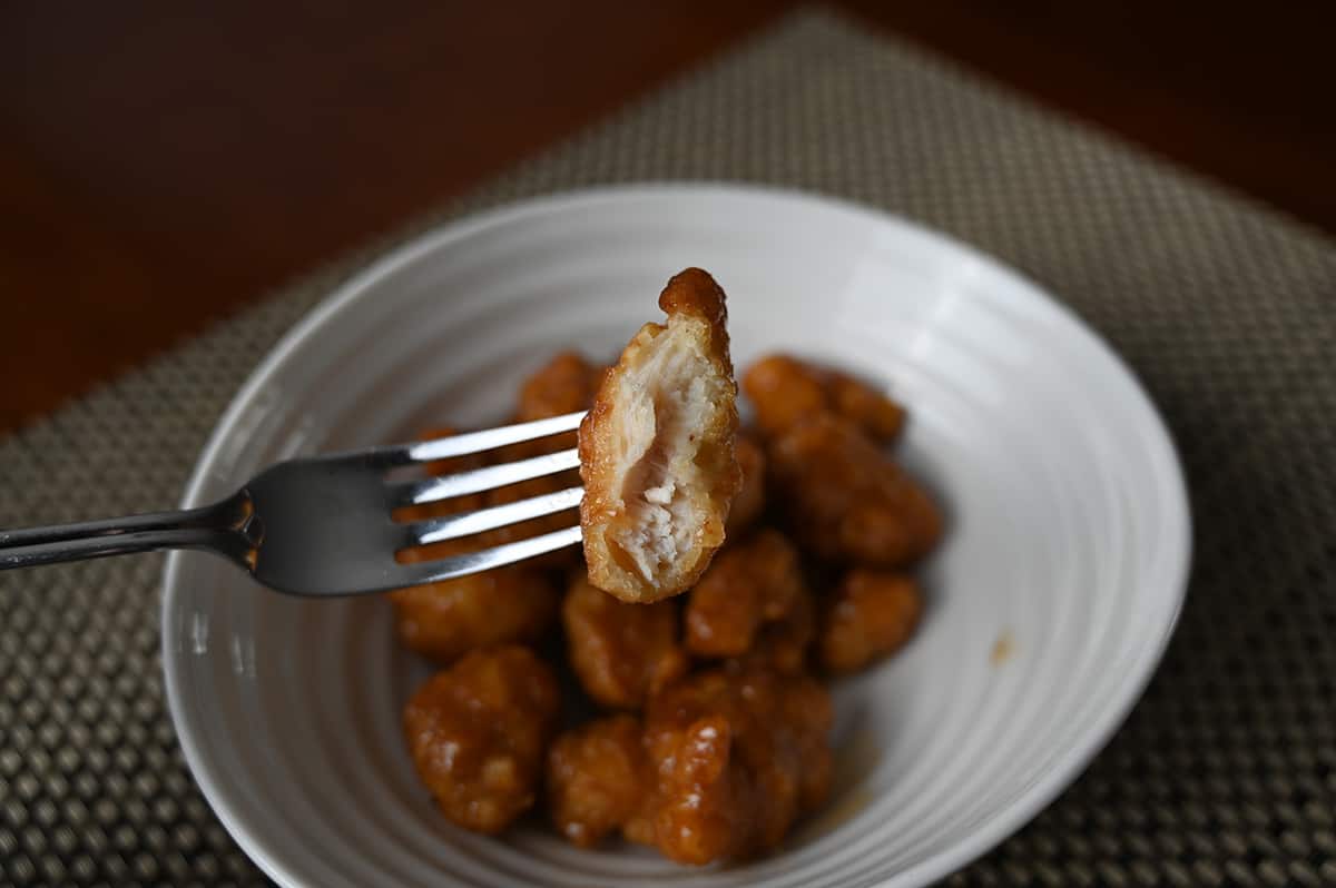 Image of a fork with one piece of chicken with a bite tsken out of it so you can see the center hovering over a bowl of orange chicken.