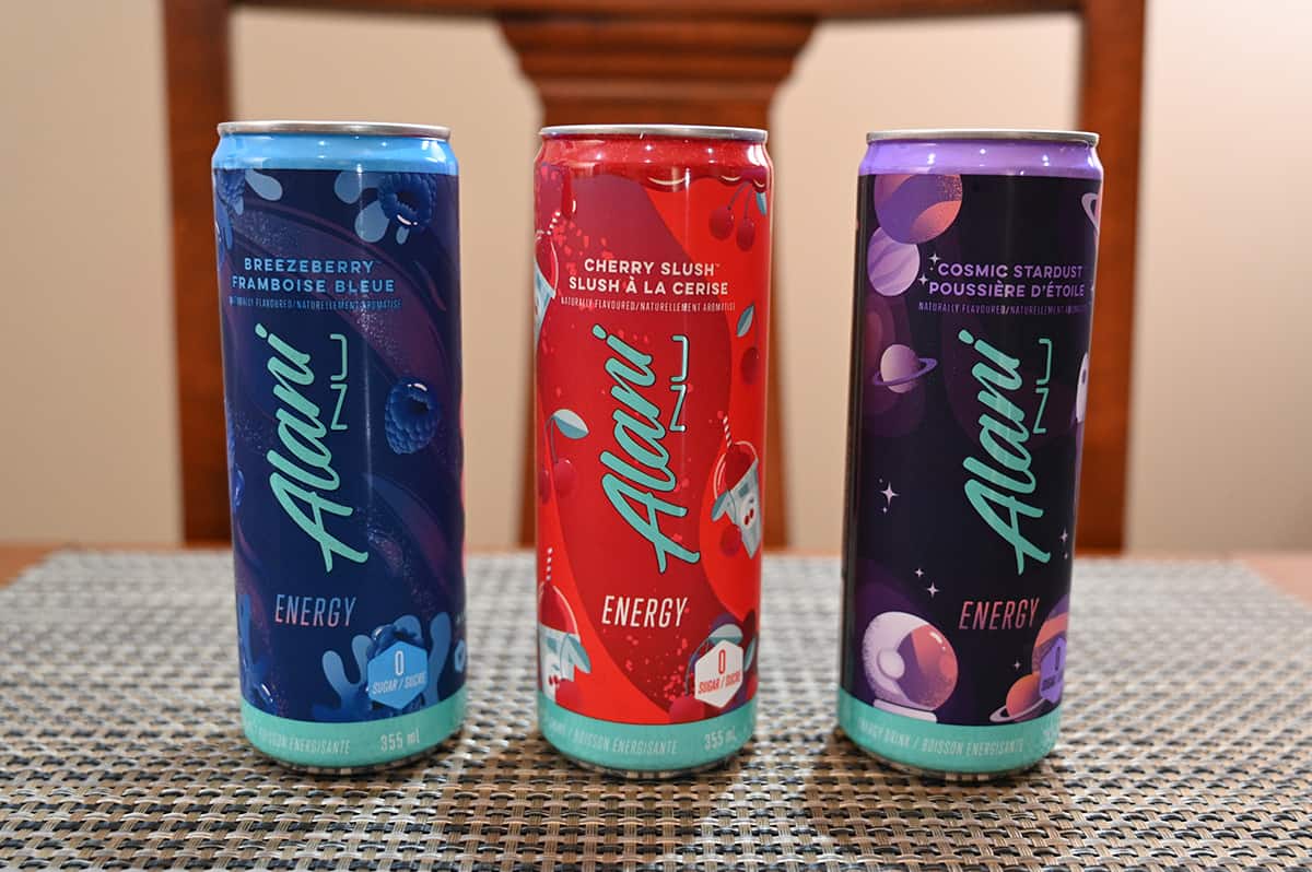 Image of three cans of energy drinks unopened on a table, there is a breezeberry, cherry slush nad cosmic stardust flavored drink.