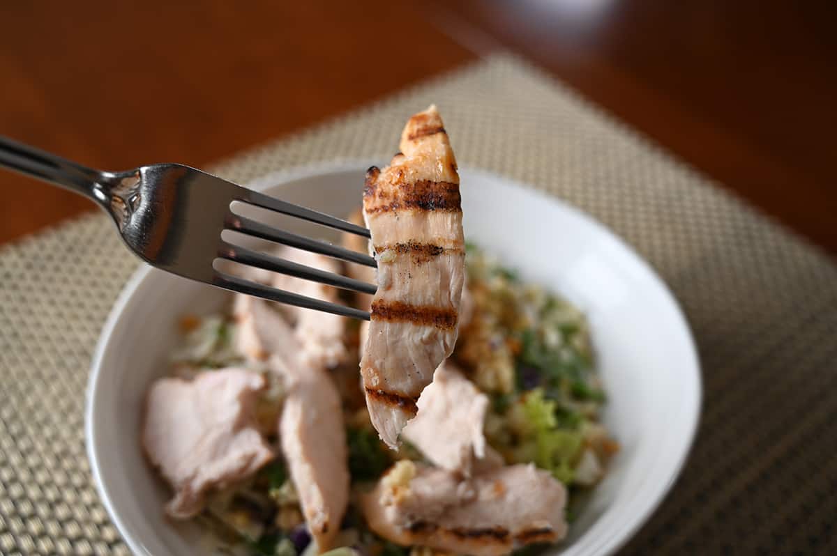 Side view image of a fork with one chicken breast strip on it with grill marks hovering over a bowl of salad with chicken.