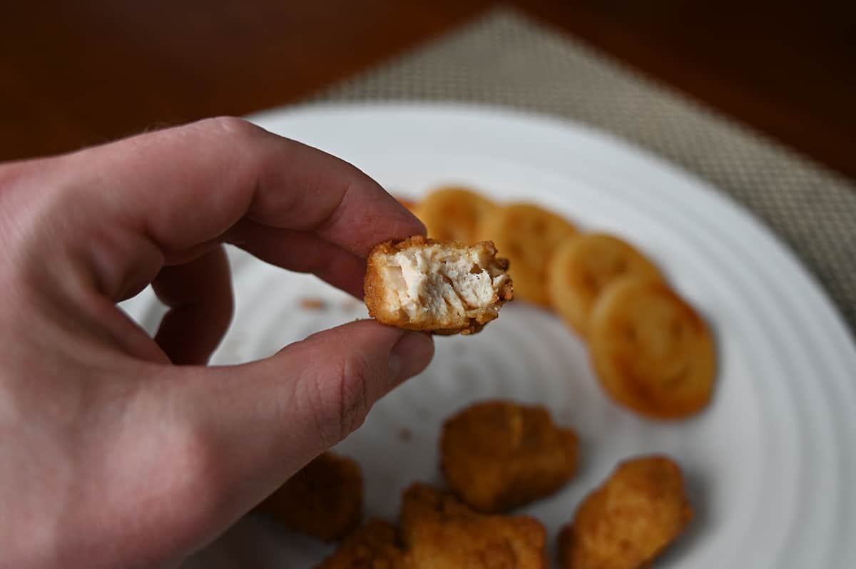 Closeup image of a hand holding one chicken breast chunk close to the camera with a bite taken out of it so you can see the middle.