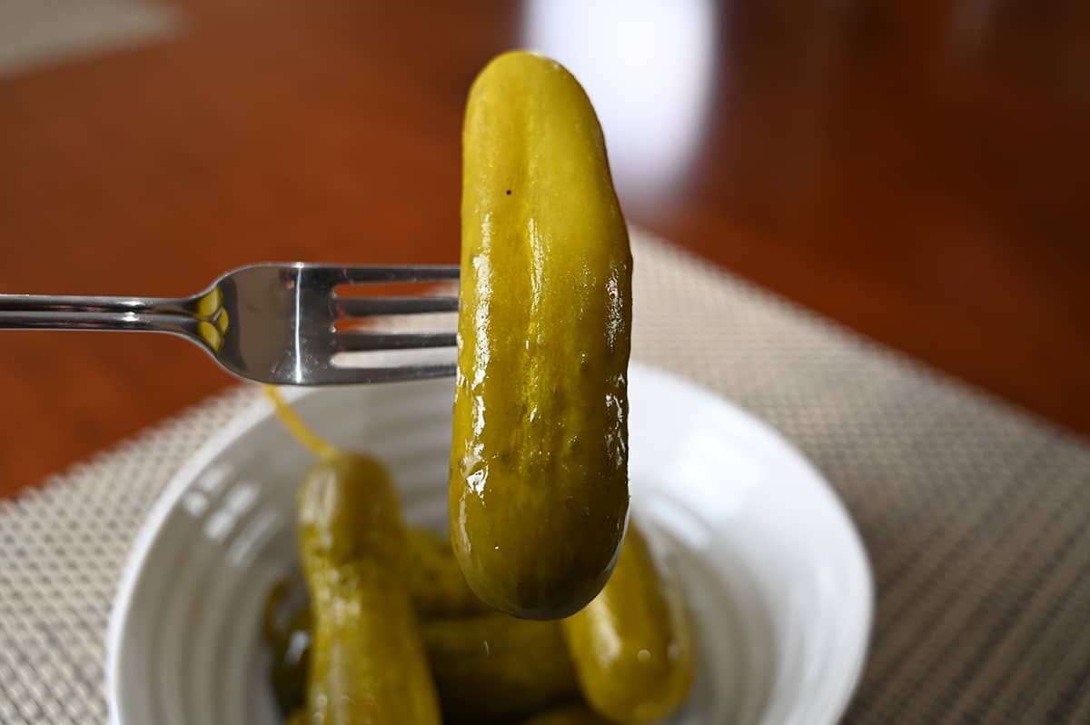 Image of a fork holding one dill pickle up close to the camera over a bowl of dill pickles beneath it.