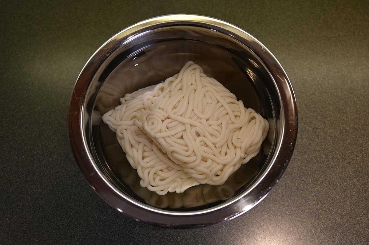 Top down image of noodles being soaked in water in a stainless steel bowl.