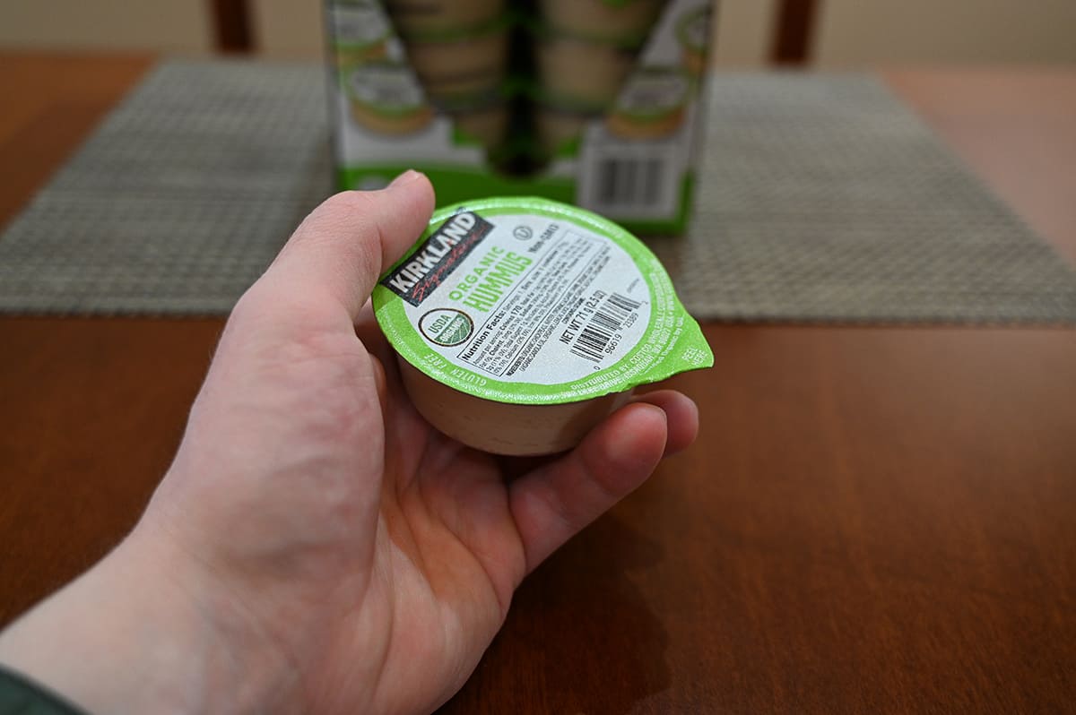 Side view image of a hand holding one individually packaged cup of hummus with a box of hummus in the background.