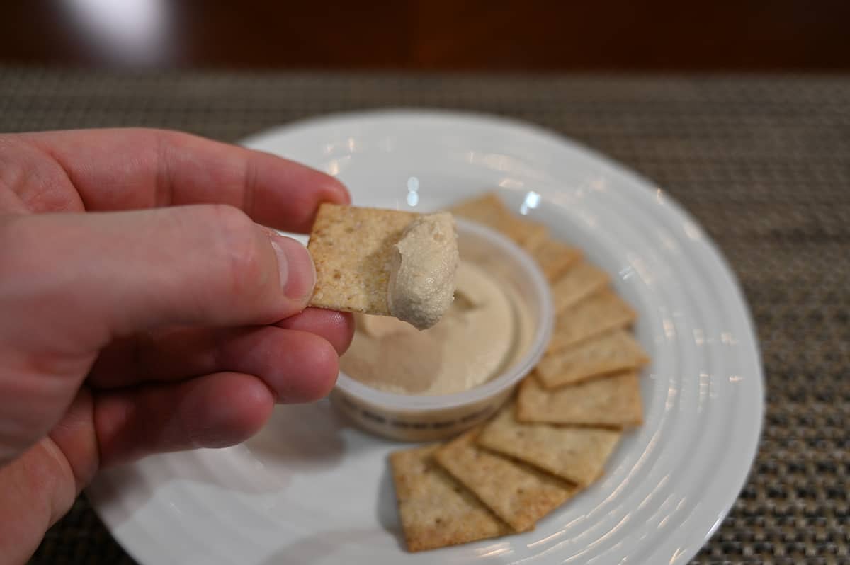 Sideview image of a hand dipping one cracker into a cup of hummus, there are crackers on the plate.
