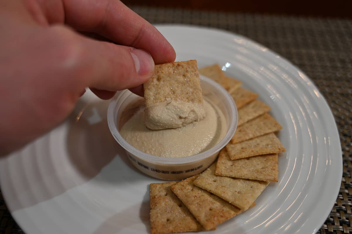 Top  down image of a hand dipping one cracker into a plastic cup of hummus with crackers surrounding the hummus.