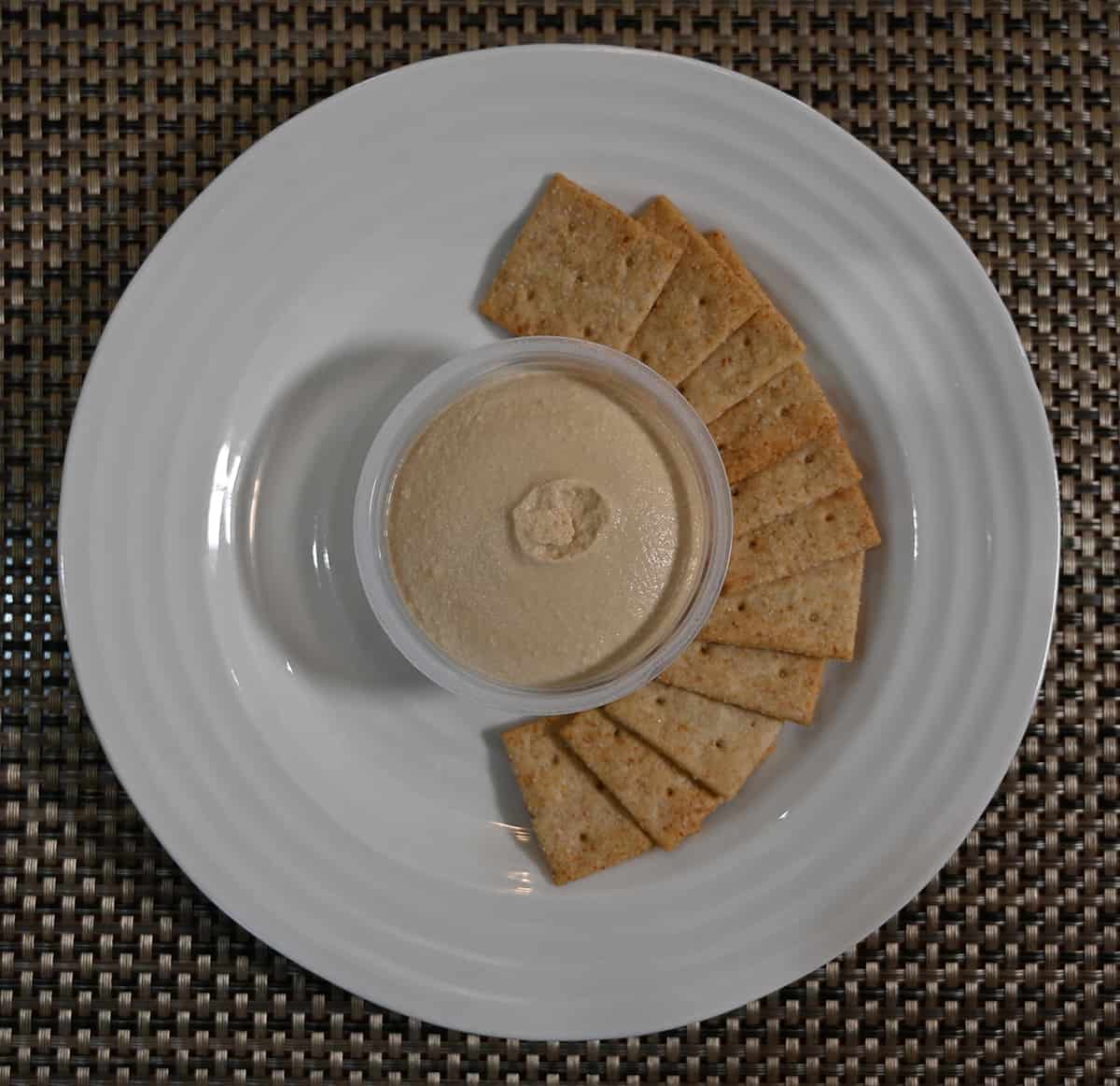 Top down image of a plate of crackers with a plastic cup of hummus in the middle of the crackers.