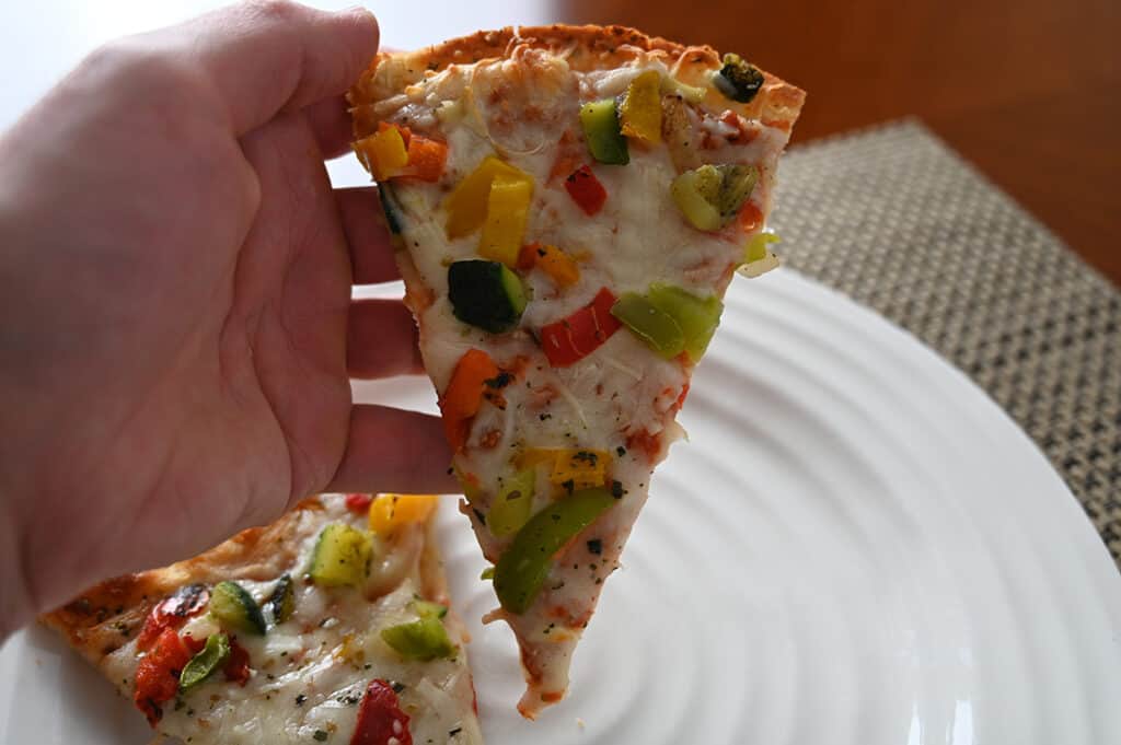 Closeup image of a hand holding one slice of cauliflower crust pizza close to the camera.