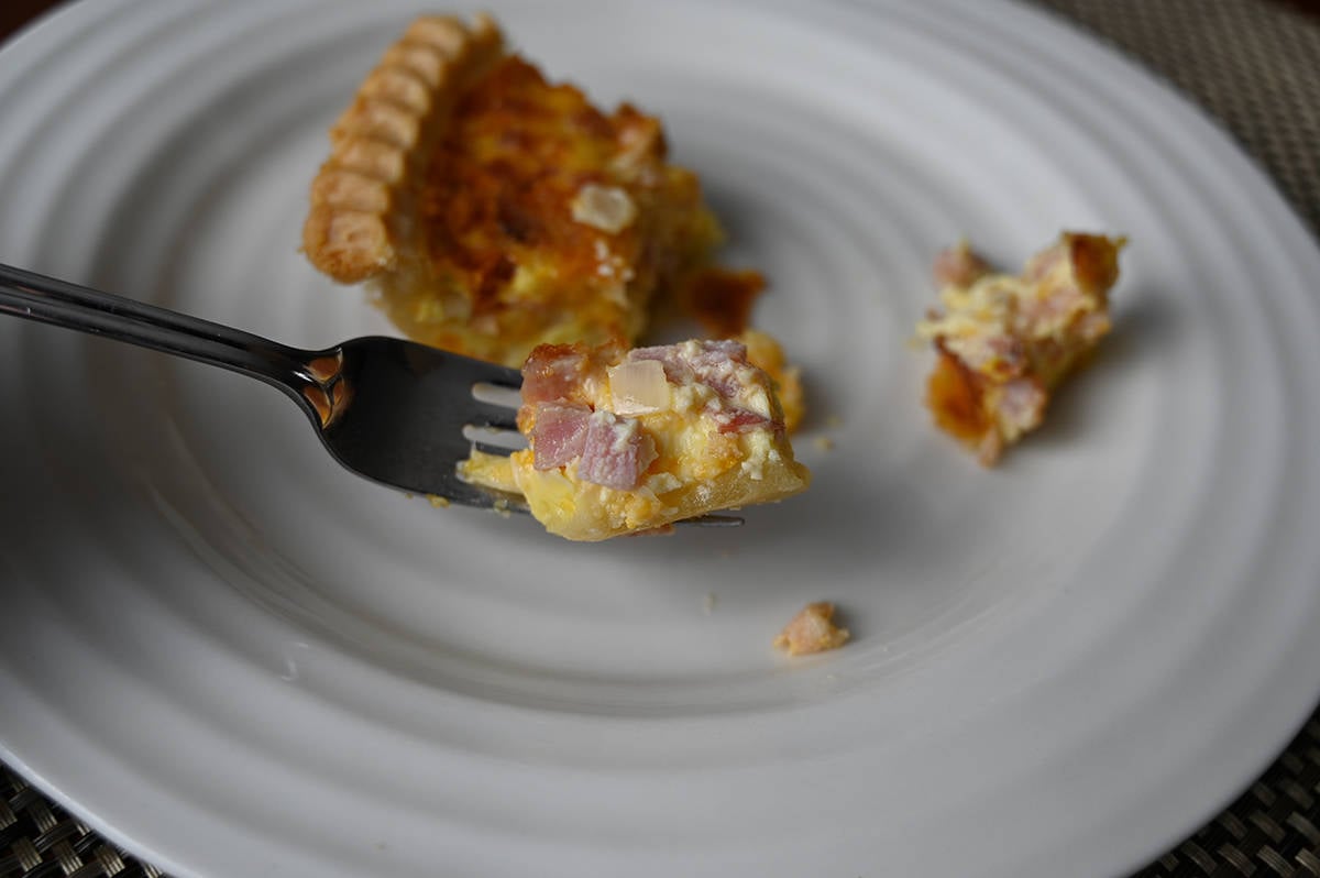 Image of a fork with a bite of quiche on it showing the ham and onions in the quiche with a piece of quiche on a plate in the background.