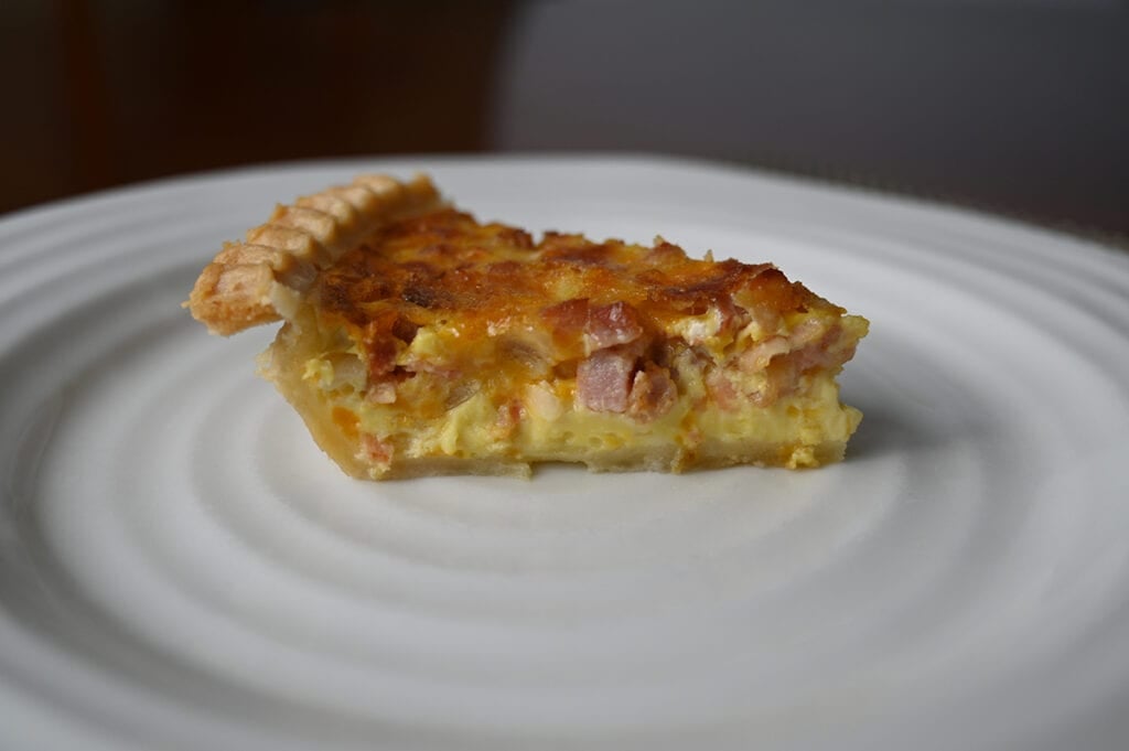 Side view image of one slice of quiche served on a white plate so you can see the filling of the quiche and the bottom crust.