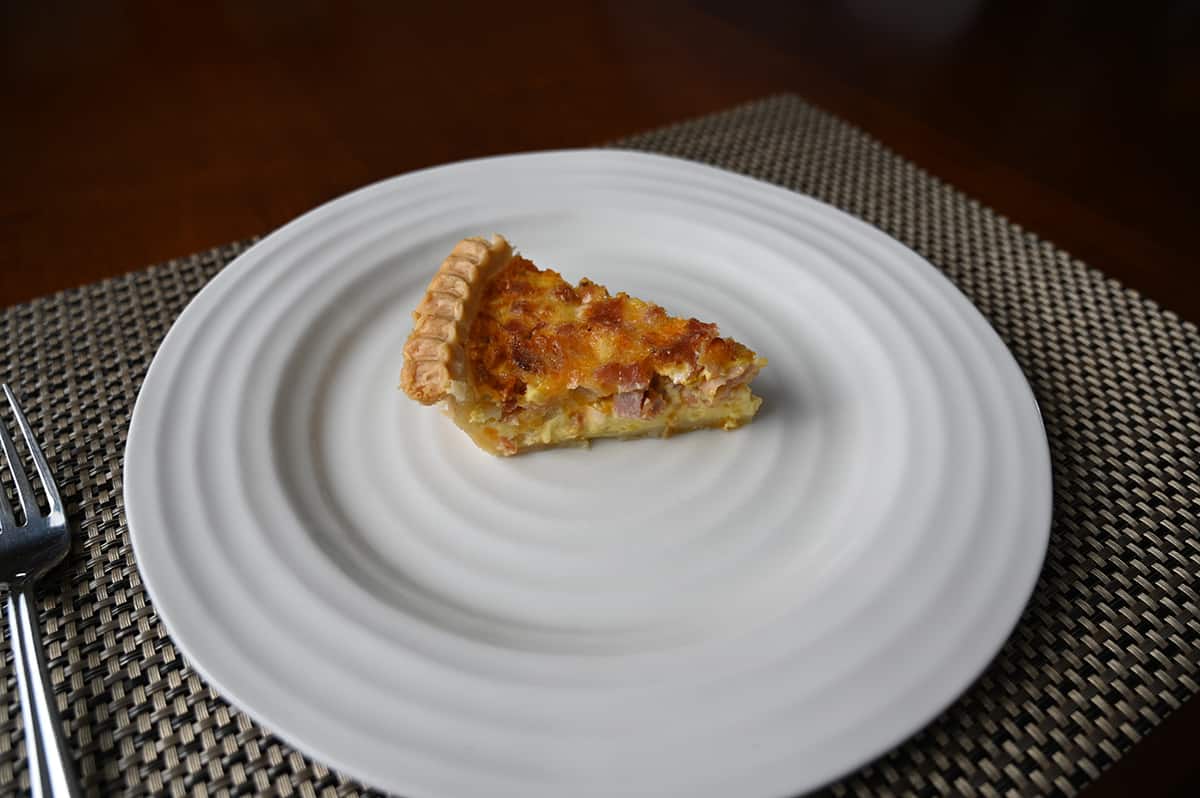 Side view top down image of one slice of quiche served on a white plate with a fork beside the plate.