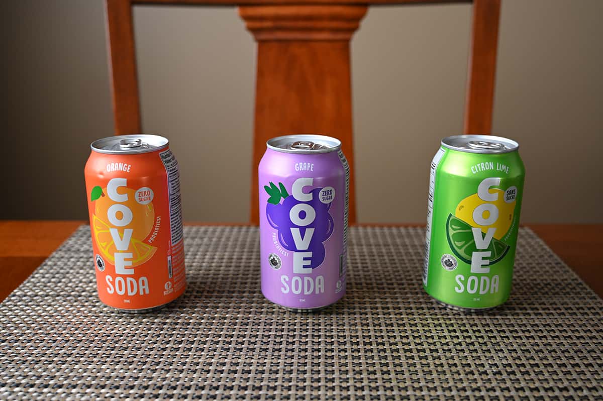 Image of three cans of soda unopened sitting on a table, there is orange, grape and lemon-lime.