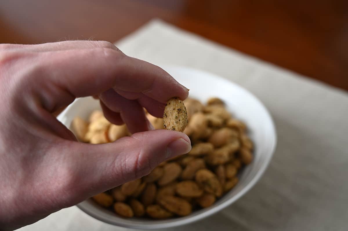Closeup image of a hand holding one seasoned almond close to the camera. In the background of the image is a bowl of almonds.