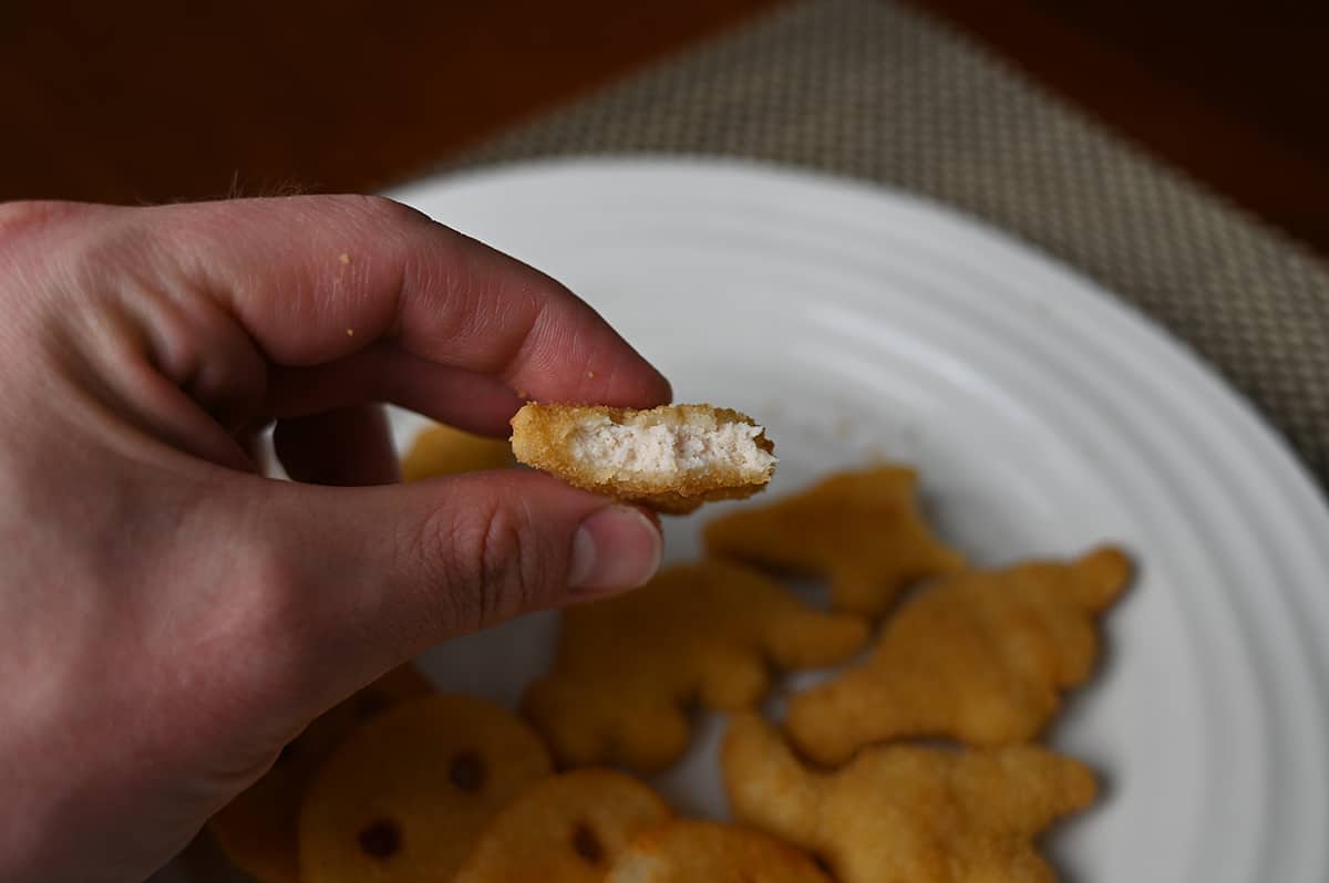 Closeup side view image of a hand holding a dino nugget with a bite taken out of it so you can see the white meat in the center.